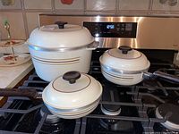 Set of three off-white cast aluminum pots with brown and gold stripes and lids, placed on stove burners