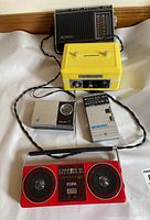 Photo showing all five items in the lot including the yellow Cashbox coin bank and four radios: Sanyo, York, GE, Craig.