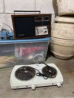 Photo shows one vintage radio and the electric travel stovetop together on a plastic container. The radio is woodgrain with a black speaker grille, multiple buttons, and tuning dial. The stovetop is white with two coil burners and a control knob in front.
