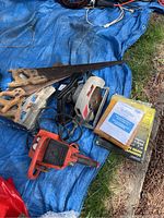Multiple saws including wooden-handled hand saws, two electric circular saws in plastic housings, and some manuals on a blue tarp outdoors.