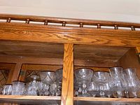Shelf displaying multiple sets of stemware including brandy snifters, dessert bowls, wine glasses, liquor glasses, and shot glasses in a wooden cabinet.