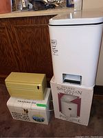 Three items shown: white metal KCO garbage bin on its original box, beige/yellow metal food wrap dispenser, and cardboard storage box with disposable touques and gloves.