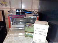 Premier toaster oven, model T610, with a saucepan on top, Kenmore electric can opener and cutter sharpener in original box, and press burger cooker with black handle, all on the kitchen counter.