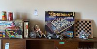 Overview of lot with boxed games and toys displayed on shelf including Mickey Mouse roller coaster, The Generals game, chess board with bagged pieces, toy car, horse carriage, unopened cans.