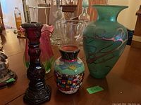 Group of various decorative vases including a green ceramic vase, colorful folk art vase, red glass vase, clear glass vase, and a pink translucent glass vase.