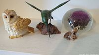 All four figurines arranged showing full lot contents: resin owl, wooden hummingbird, red rose tea fawn with chip, and glass paperweight with Scotch thistle flower.