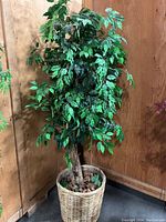 Large artificial tree in a wicker pot against a wooden wall with dense dark green leaves and visible base with artificial soil and bark.