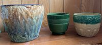 Three ceramic plant pots on a wood surface against a wood-paneled background: a large multicolor textured pot, a medium green pot with raised pattern, and a cream pot with teal floral decoration.