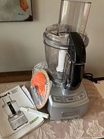 Full view of the Cuisinart food processor unit, showing the base, bowl, lid, and black handle. Also includes instruction manual, plastic pusher, and metal blade sealed in plastic on the table beside it.
