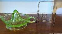 Photo showing green glass citrus juicer with a handle and metal holder with wire handle and slots side by side on table.