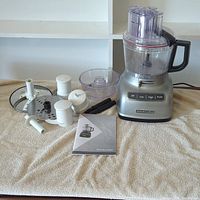 Full lot showing KitchenAid food processor base, clear plastic bowl and lid, white plastic attachments, and manual on a beige towel.
