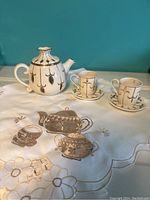 White ceramic teapot-shaped votive candle holder with two matching cup-shaped votive holders on embroidered tablecloth