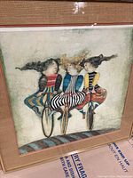Full view of the framed artwork showing three women riding bicycles in colorful striped outfits against a muted textured background, framed in a gold frame with a textured mat.