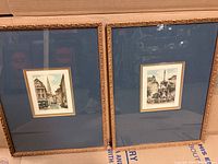 Two framed and matted watercolour artworks with blue mats and gold ornate frames, placed side by side showing detailed urban architectural scenes.