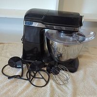 Front left profile of black Sunbeam countertop electric mixer with three attachments displayed: dough hook, flat beater, and wire whip, and stainless steel bowl with splash guard.