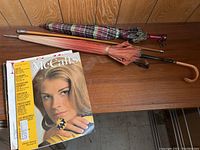 Three umbrellas and vintage magazines placed on wooden surface, showing overall lot content including pink fabric umbrella, plaid umbrella, and magazines.