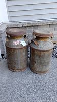 Pair of two vintage metal milk cans standing side by side outdoors showing overall size and condition.