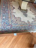 Top-down view of rug partially under glass table showing floral pattern and blue border on hardwood floor