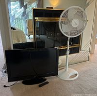 Photo of black LG flat-screen TV, remote, and white oscillating fan on carpeted floor
