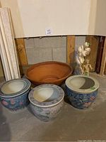 Photo showing three Chinese ceramic pots with floral and botanical paint details, a large terra-cotta pot behind them, and two small garden statues to the side.