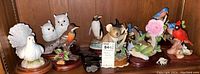 Porcelain and ceramic bird and animal figurines displayed together on shelf. Variety of species including owls, pigeon, penguin, mice, robins, cardinal, and bird with flower. One piece noted musical.