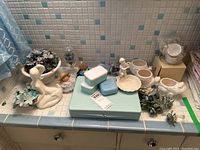 Wide view of bathroom accessories on tiled counter showing soap dishes, planters, faux flowers, and figurines.