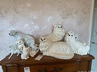 Photo showing ceramic animal figurines on a wooden surface against floral wallpaper. Includes large Royal Dux polar bear and multiple white cat figurines, as well as unicorns.