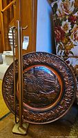 Photo showing pictorial hammered copper wall plate propped vertically with brass fireplace tools beside it.
