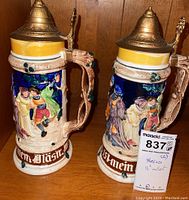 Pair of two German style lidded steins with ornate relief artwork, yellow and blue glaze, and pewter lids.