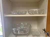 Multiple glass and crystal bowls displayed inside a shelf, showing various sized footed bowls including divided and scalloped edged pieces with clear glass and etched starburst designs.