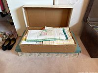 Box containing folded kitchen linens including embroidered and crocheted items with visible colorful hand embroidery and vintage towels