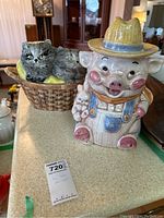 Pig cookie jar and ceramic basket bowl with cats on countertop with identification tag.