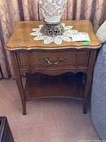 Photo of one wooden nightstand showing single drawer with decorative handle, lower open shelf, and curved edges on sides and legs.