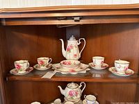 Full set displayed on shelf including coffee pot, serving dish, four teacups and saucers arranged in front, and three bowls.