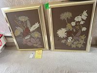 Two framed floral artworks by Jillian Field displayed side by side on carpeted floor. Each frame is gold-tone measuring approximately 22x28 inches containing brown-background floral art.
