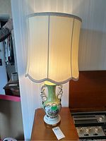 Table lamp standing on a wooden surface, showing the full lamp lit, with ceramic decorated base and cream-colored scalloped fabric shade.
