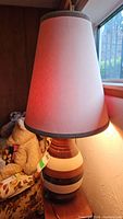 Overall view of the lamp turned on, showing the vertical tapered beige fabric shade with brown trim and glowing light inside, the base made of shiny pottery with broad horizontal stripes in shades of brown and cream.