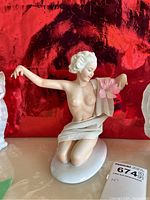 Full view of porcelain figurine of kneeling lady on oval base against red background with an auction tag.