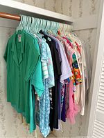 Full rack view of vintage women's blouses and tops showing a variety of colors and patterns hung on blue plastic hangers against floral wallpaper.