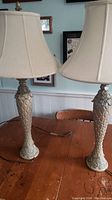 Full view of two vintage table lamps with beige fabric shades and tall decorative resin bases on a wooden table.