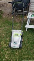 Front and top angled view of the electric lawn mower showing white and green plastic body, black metal handle with coiled power cord, and wheels on grass.