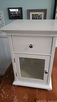 Front view of white wooden side table showing single drawer with metal knob and glass door cabinet below with metal knob, placed on wooden floor against a wall with picture frames.