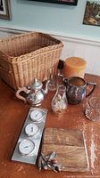 Photo showing wicker basket, silver teapot, glassware, candle, barometer and cutting board on a wood table.