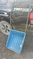 Full view of a blue metal snow scoop with a black tubular handle, positioned against the side of a black vehicle.
