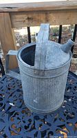 Photo of vintage galvanized tin watering container with arched handle and angled spout on black decorative metal table near wooden railing outside.