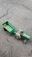 Green metal Tonka tractor connected to green metal wagon, photographed outdoors on cracked pavement.