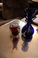 Photo showing both glass pieces displayed on a metal surface, with the cobalt blue vase and the red and clear glass snail sculpture side by side.