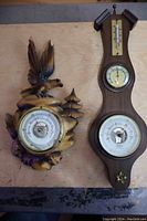 Two vintage wooden case barometers side by side on wooden surface, one Black Forest style with carved bird and tree, other Fisher barometer with thermometer above dial
