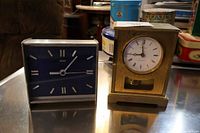 Two metal clocks made in West Germany on a reflective surface, one brass with pendulum and one with blue face