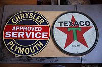 Both Chrysler Plymouth and Texaco round metal automotive signs placed side by side on a table.